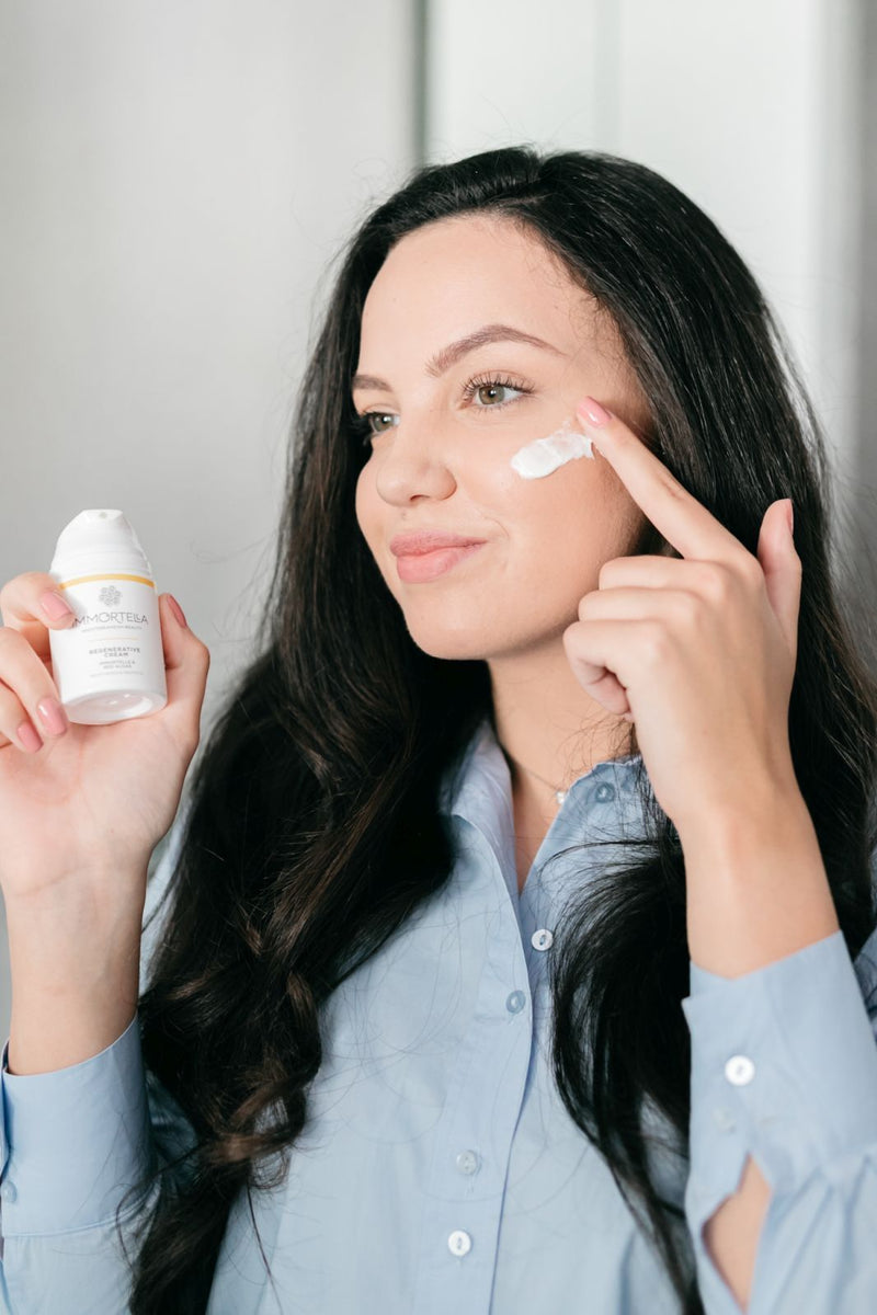 Beautiful young women with long dark hair is applying immortelle cream with fingers on her face