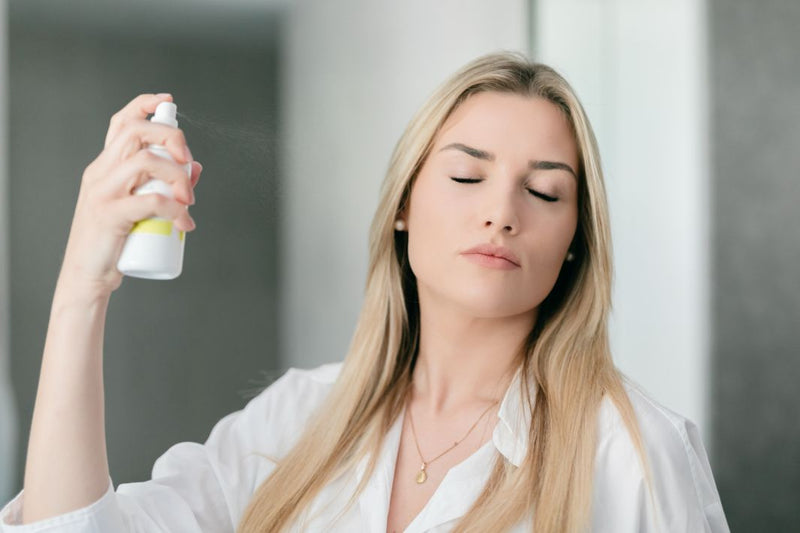 An attractive young woman, with long blond hair is spraying her face with hydrating immortelle facial mist toner made of immortelle floral water.
