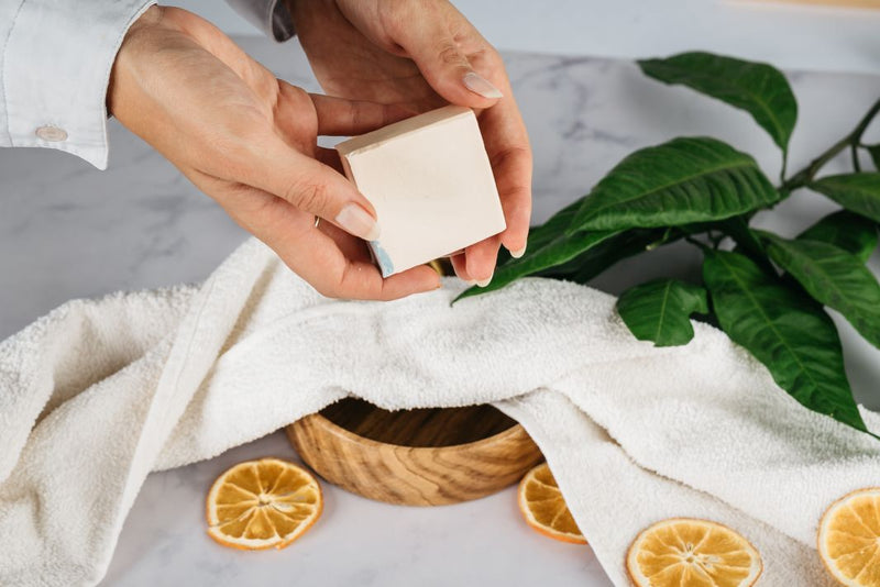  Close up of women’s hands holding natural handmade soap bar. In the background of the picture is wooden bowl, half covered with white towel. The bowl is on marble table. There are couple of orange slices on the table and on the towel as well as leafs of orange tree.