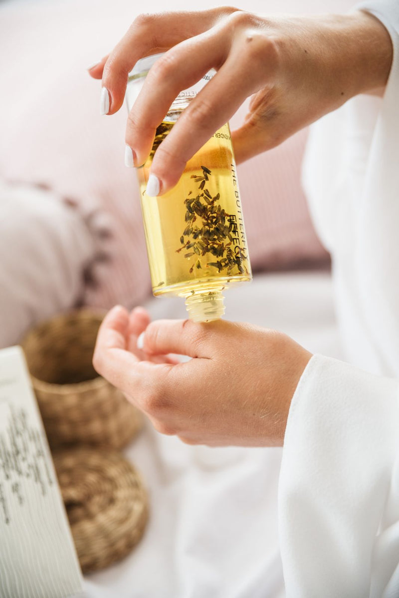 Close up of young women’s hands pouring lavender body oil in her hands. Glass bottle of natural body oil infused with dried lavender flowers.