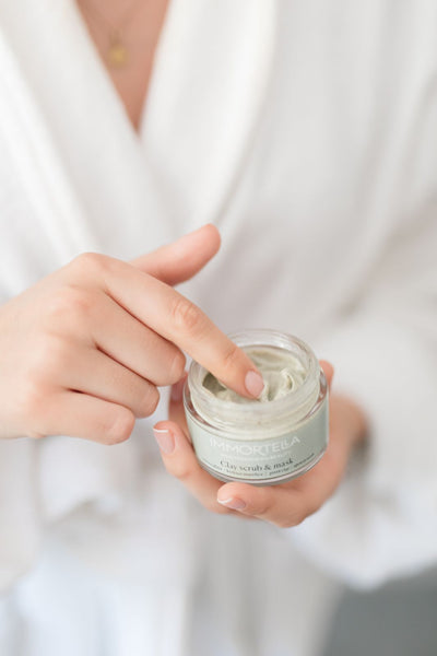 Close up of women in bathroom robe, holding jar of natural clay facial scrub and mask. She is trying the texture of face mask with her fingers, before applying it on her face.