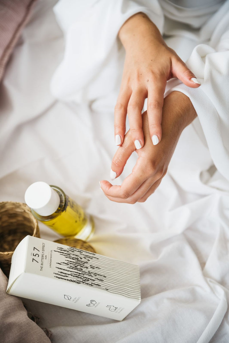 Close up of young women in silk bathrobe applying natural handmade lavender body oil.  Beside her is glass bottle of natural oil and its white eco-friendly packaging. On the packaging is beautiful sketch of lavender flowers.