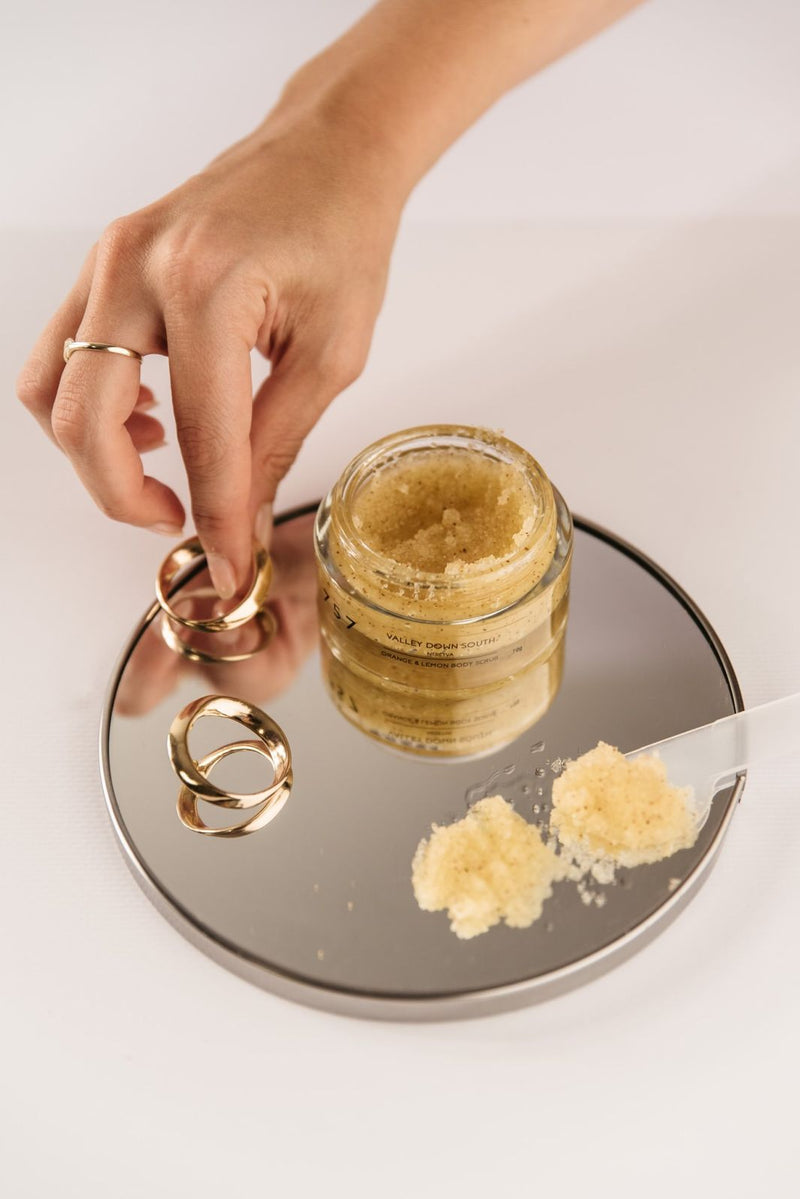 Glass jar of body scrub made of sea salt, orange and lemon on glass board.  On the glass board are couple of golden rings and women’s finger trying to pick up one of the ring and smear of citrus scrub.