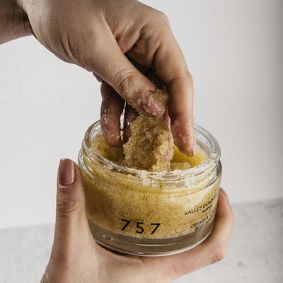 Close up of women’s hands. With one hand she is holding natural handmade body scrub made of orange lemon and sea salt. Her other hand is jar, she is tying the structure of the body scrub with her fingers.