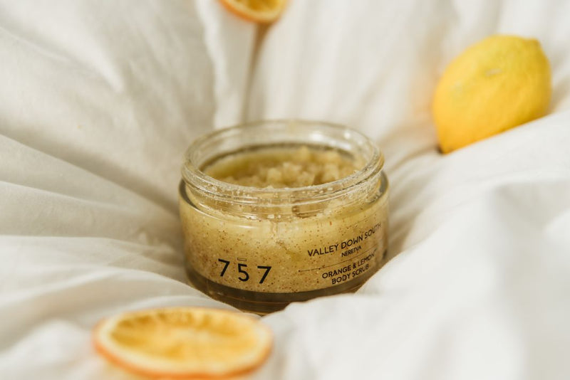 Close up of open natural handmade citrus and sea slat body scrub on the bed on the quilt. Around the lemon and orange body scrub is one whole lemon and couple of lemon slices.