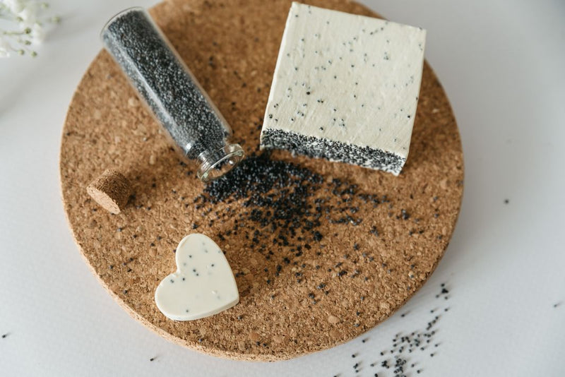 Close up of two soaps bar, one of them is small and heart shaped. The natural handmade soaps are lying on the cork board next to the little glass bottle of poppy seeds which is open and little seeds are scattered on the cork board. Soap is made of extra virgin Croatian olive oil, and pure mint, clove and cinnamon essential oils.