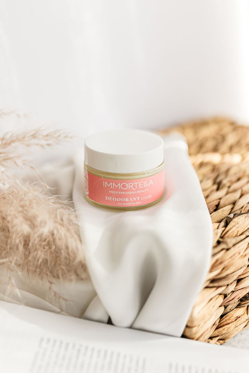 Glass of natural deodorant cream standing on wicker basket, one half of basket is wrapped in beautiful silk fabric, beside product is decorative pampas grass.