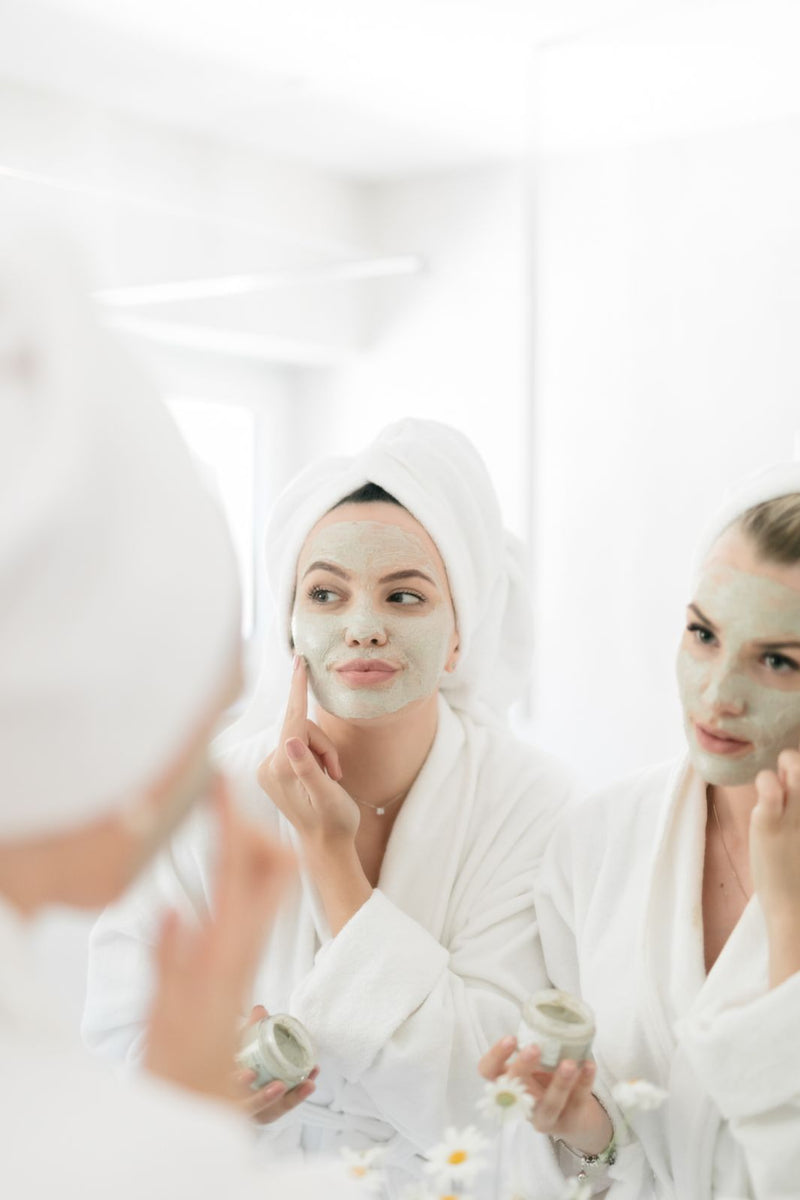 Reflection of two beautiful young women in bathroom mirror, applying natural clay and scrub face mask. 